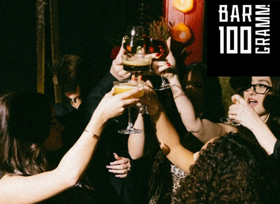 Eine Gruppe von Menschen erhebt in einer schwach beleuchteten Berliner Bar die Gläser und stößt an. In der Ecke ist ein Schild mit der Aufschrift BAR 100 GRAMM" zu sehen.
